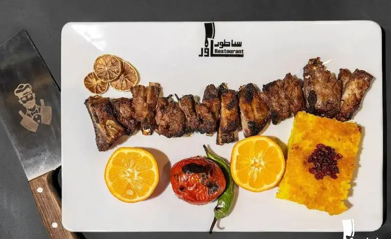 دنده کباب آبدار و خوشمزه رستوران ساطور، سرو شده با برنج زعفرانی و زرشک، پرتقال تازه و گوجه کبابی، غذای اصیل ایرانی.