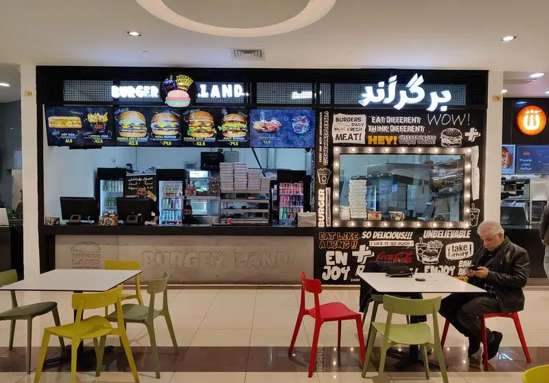 تصویر داخلی فست فود برگر لند با منوی دیجیتال بزرگ برگرها، کانتر سفارش، پرسنل، و چیدمان میز و صندلی‌های رنگی برای مشتریان.