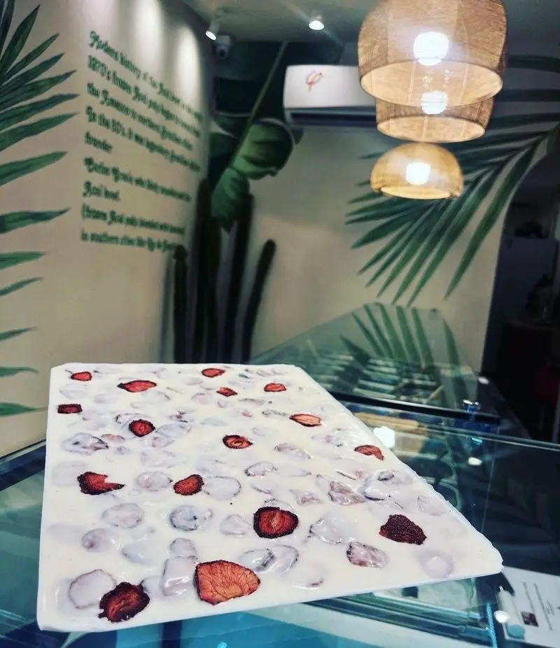 دسر بزرگ و خنک آسایی کاسه روح با توت فرنگی خشک و تکه های شفاف روی پیشخوان شیشه ای کافه. دیوار با نقوش برگ نخل و نورپردازی گرم.