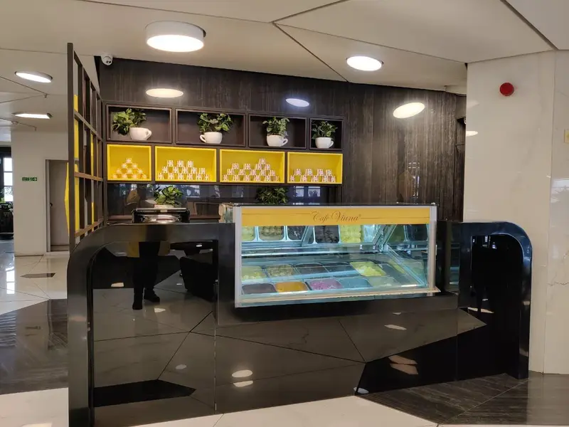 پیشخوان مدرن کافه ویونا فود کورت رویال با نمایشگر بستنی‌های رنگارنگ، دکوراسیون شیک با قفسه‌های زرد و گیاهان تزئینی