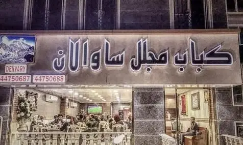 نمای بیرونی و تابلوی روشن رستوران کباب مجلل ساوالان، همراه با شماره های تماس دلیوری و فضای داخلی پر مشتری.