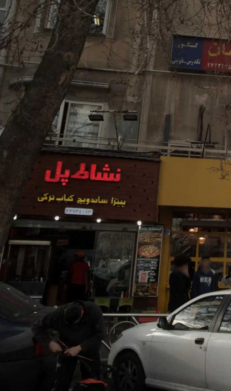 فست فود نشاط پل: نمای خیابانی مغازه با تابلوهای قرمز نئونی، ارائه دهنده پیتزا، ساندویچ و کباب ترکی.