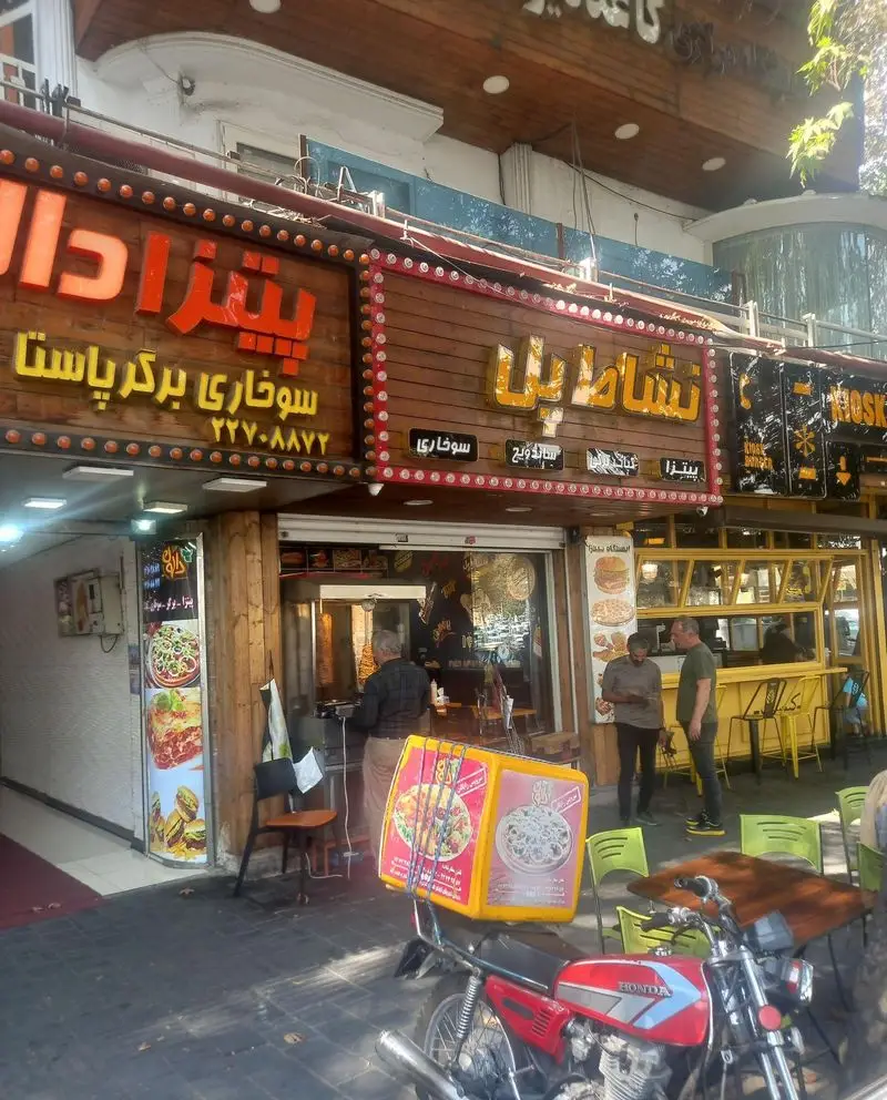 فست فود نشاط پل با نمای چوبی و تابلوهای پیتزا، برگر، سوخاری، ساندویچ و پاستا. میز و صندلی بیرونی و موتور بیرون‌بر در تصویر.