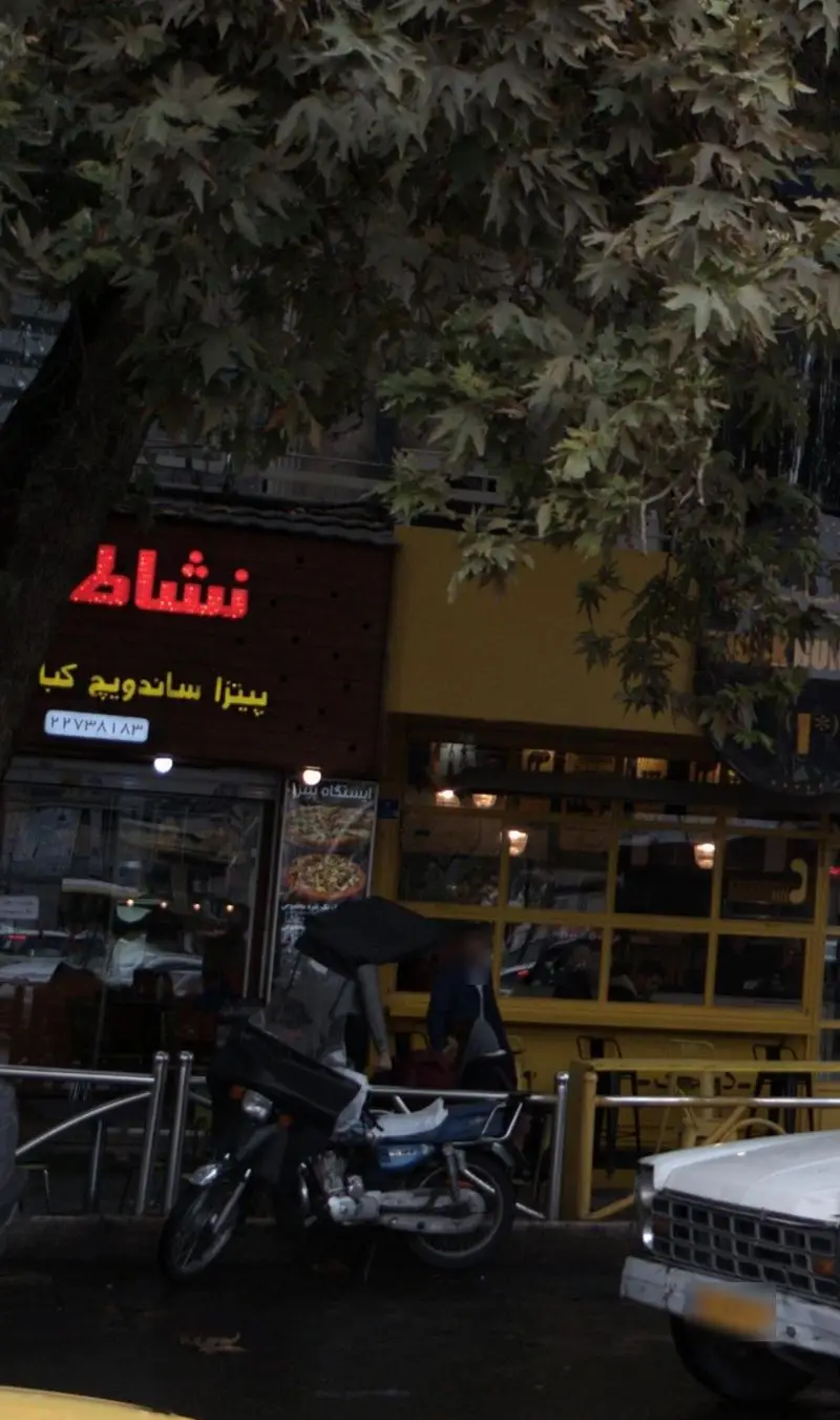 نمای بیرونی فست فود نشاط پل ارائه دهنده پیتزا، ساندویچ و کباب در خیابان، با موتورسیکلت تحویل غذا و درختان سبز.