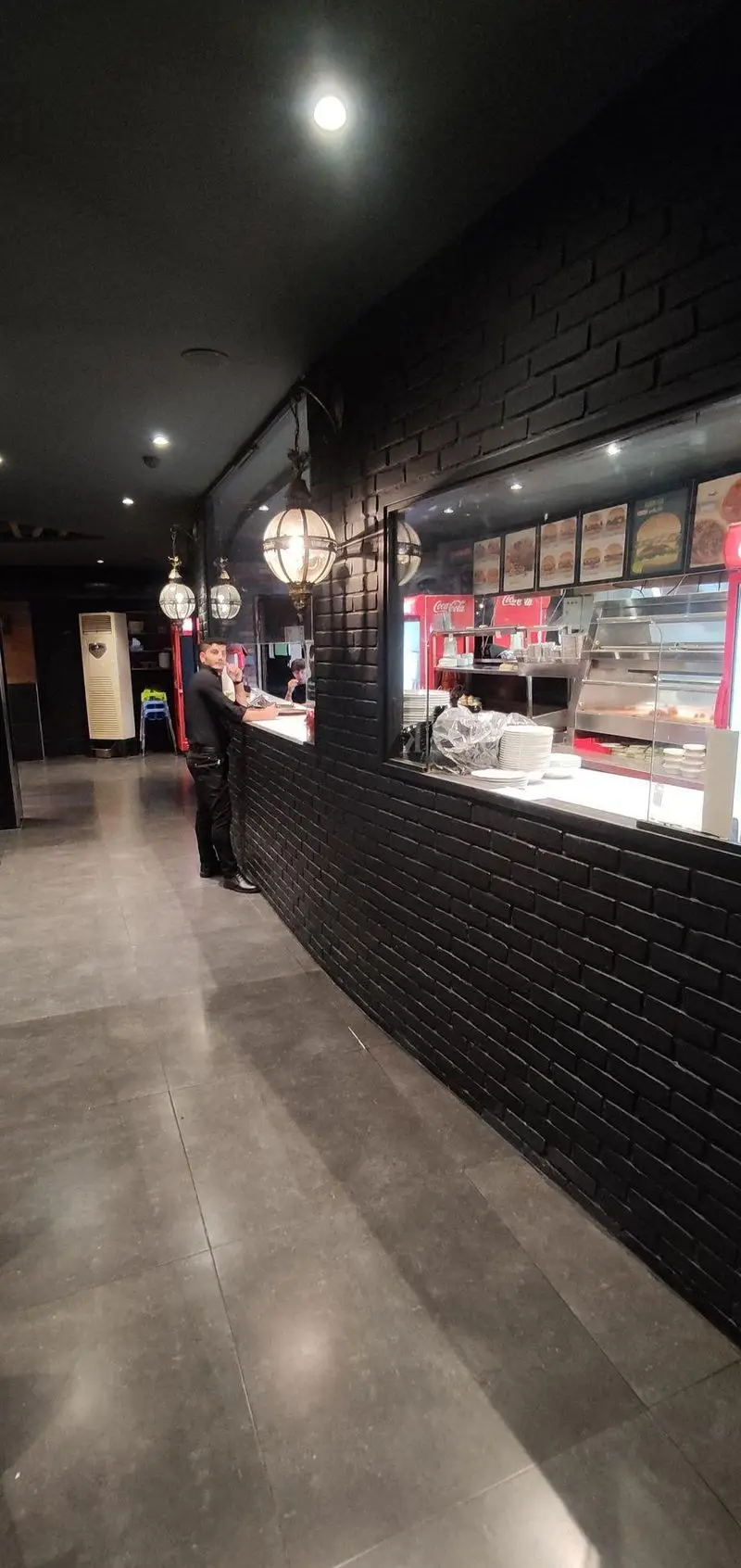 نمایی از فضای داخلی رستوران ایتالیایی مریخ، شامل کانتر سفارش غذا، دیوار آجری مشکی، لوسترهای تزئینی و یخچال‌های نوشیدنی با منوی کامل.