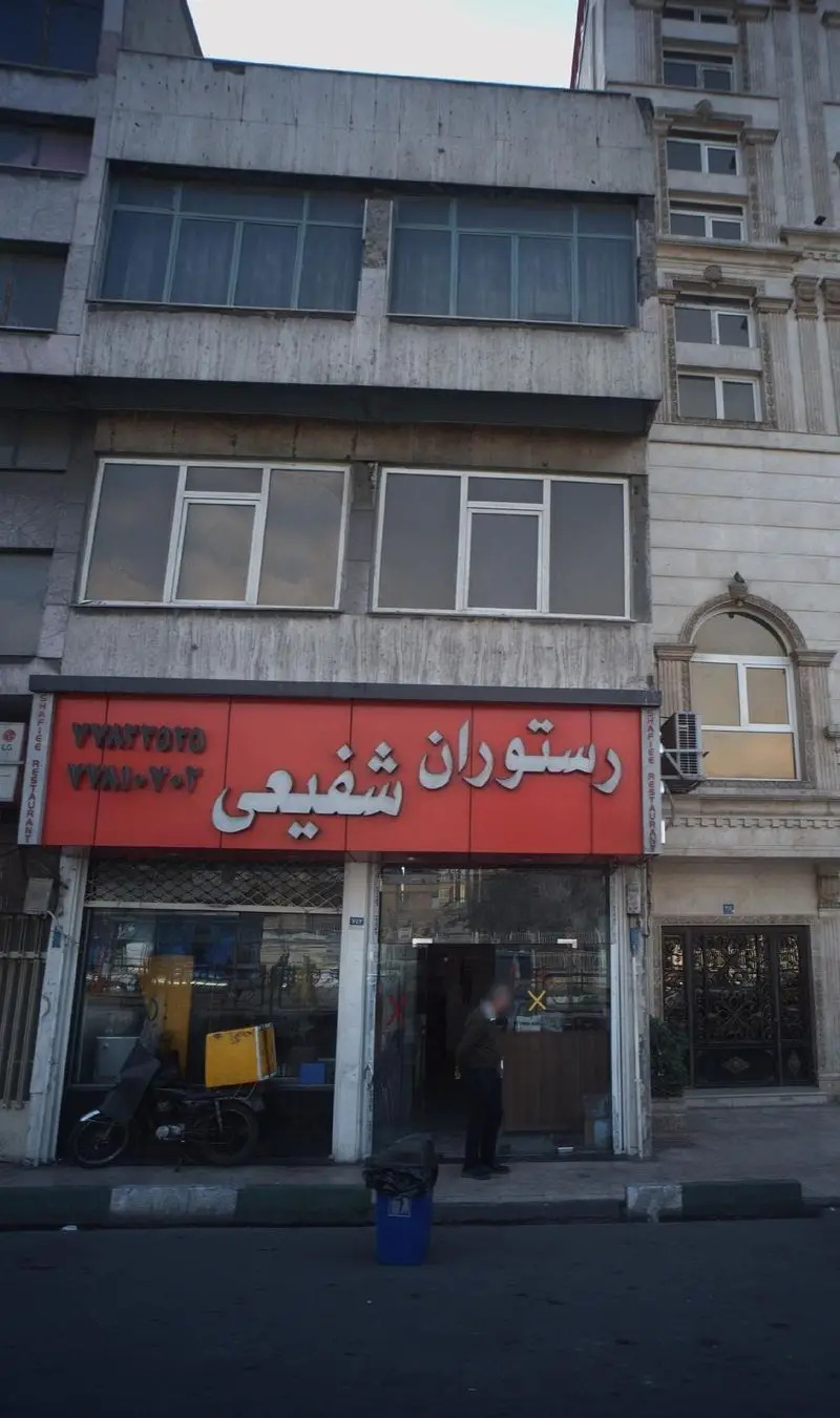نمای بیرونی رستوران شفیعی با تابلوی قرمز و سفید، ساختمان شهری با پنجره‌های متعدد و موتورسیکلت تحویل غذا در جلوی آن.