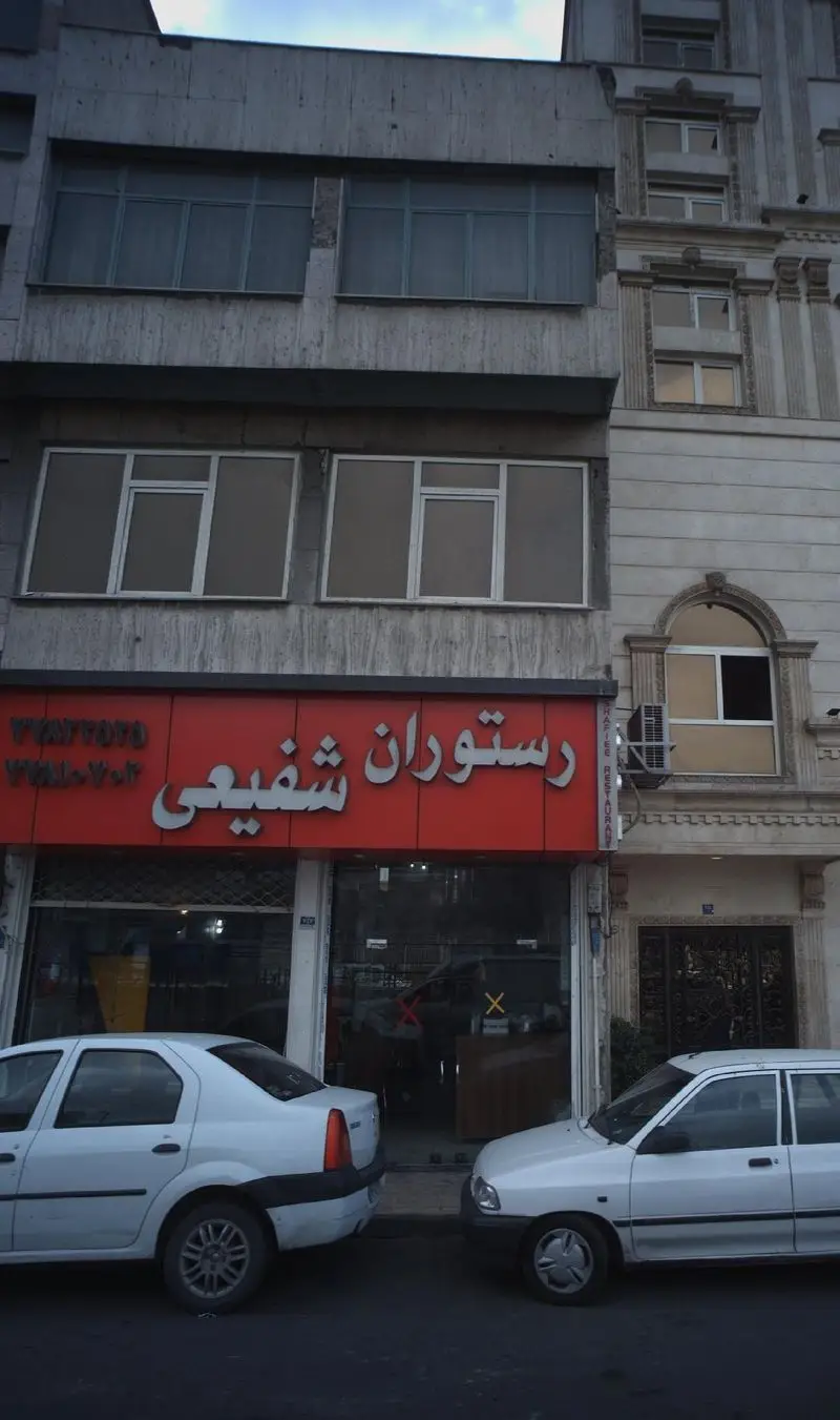 نمای خارجی رستوران شفیعی با تابلوی قرمز بزرگ، خودروهای سفید پارک شده در مقابل و ساختمان چند طبقه در خیابان شهری.
