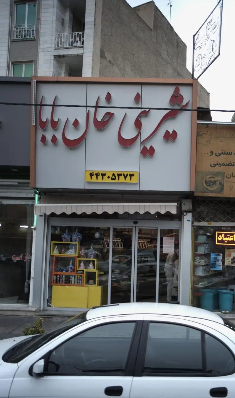 تصویر نمای بیرونی مغازه شیرینی خان بابا، یک قنادی با تابلوی قرمز رنگ و ویترین جذاب حاوی انواع شیرینی و دسر. شماره تماس: ۴۴۳۵۲۷۳.