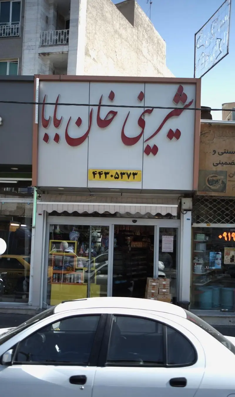 تصویر نمای بیرونی قنادی شیرینی خان بابا با تابلو قرمز رنگ و شماره تماس ۴۴۳۰۵۳۷۳ در یک روز آفتابی.