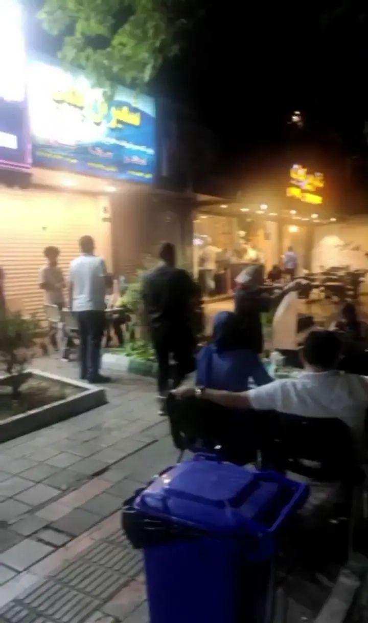 نمای شبانه رستوران تیکن کباب. مشتریان در حال غذا خوردن و تردد در پیاده‌رو. تابلوی روشن مغازه، فضای پر جنب‌وجوش و سطل زباله آبی.