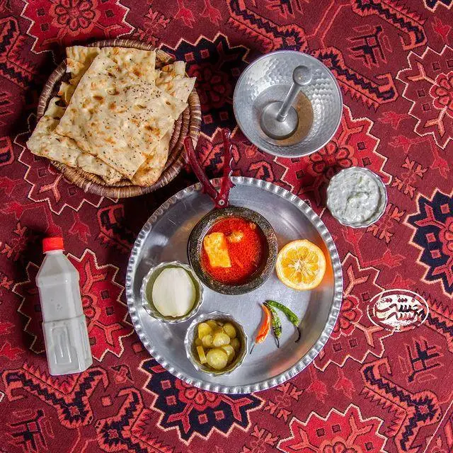 نمای بالا از سفره سنتی کافه رستوران خشت بهشت با غذای اصیل دیزی، نان تازه، دوغ، ترشی، ماست و خیار روی قالی قرمز.