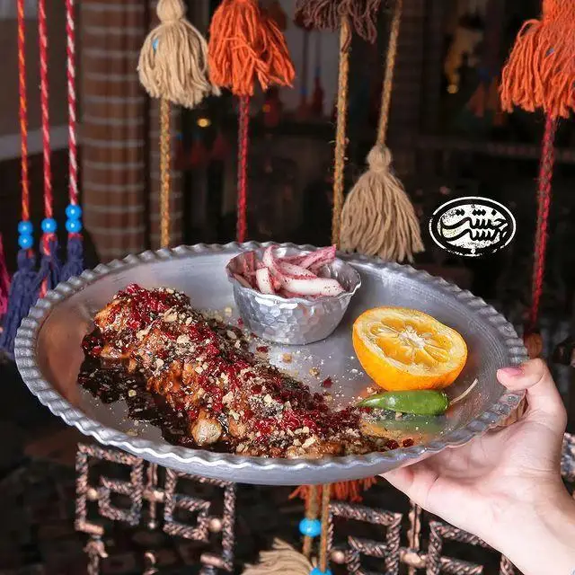 نمای نزدیک از غذای سنتی ماهی یا خوراک ویژه در کافه رستوران خشت بهشت، سرو شده با زرشک، مغز آجیل، پیاز و پرتقال در بشقاب فلزی.