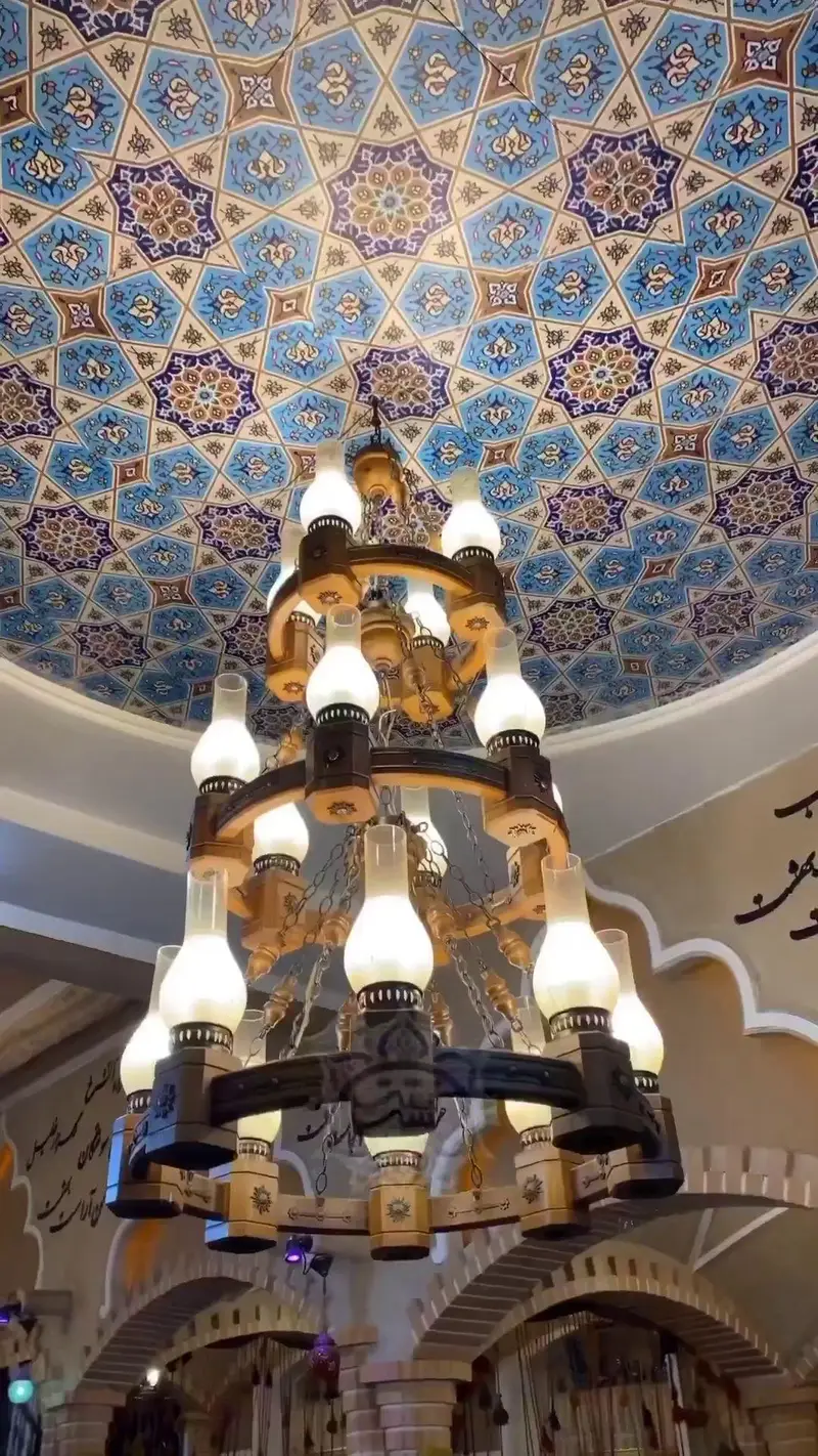 لوستر چوبی سنتی باشکوه در کافه رستوران خشت بهشت، زیر سقف کاشی‌کاری خیره‌کننده با نقوش هندسی و رنگ‌های آبی و طلایی، فضای اصیل و دلنشین ایرانی را به نمایش می‌گذارد.