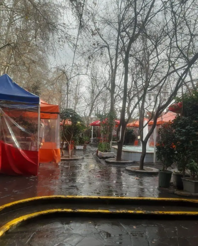 باغچه خانوادگی آبشار، کافه و رستوران فضای باز در روزی بارانی پاییزی با چادرهای رنگارنگ و محیطی دلنشین برای پذیرایی.