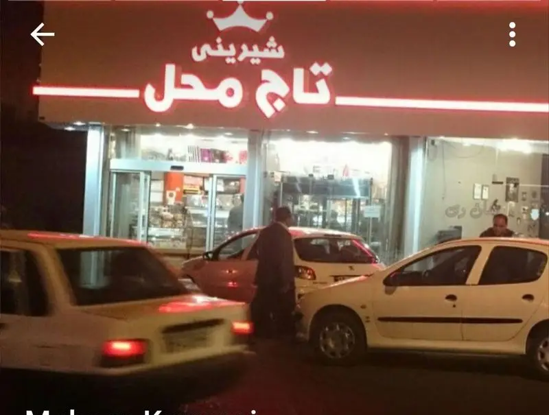 عکس نمای بیرونی شبانه قنادی و شیرینی‌فروشی تاج محل با تابلوی نئون روشن، ویترین پر از شیرینی و چند خودرو جلوی فروشگاه.
