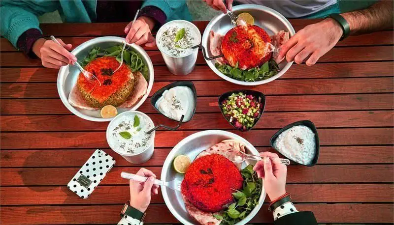 تصویری از مشتریان در پلاتین فود گاردن که دور یک میز چوبی نشسته و از غذاهای خوشمزه شامل دیس‌های رنگی، سبزیجات تازه، ماست و سالاد لذت می‌برند.