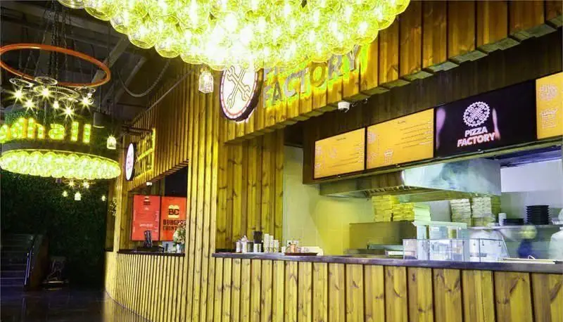 تصویر فضای داخلی پلاتین فود گاردن با پیشخوان‌های چوبی، نورپردازی مدرن سبز و زرد، تابلوی پیتزا فکتوری و منطقه فود هال.