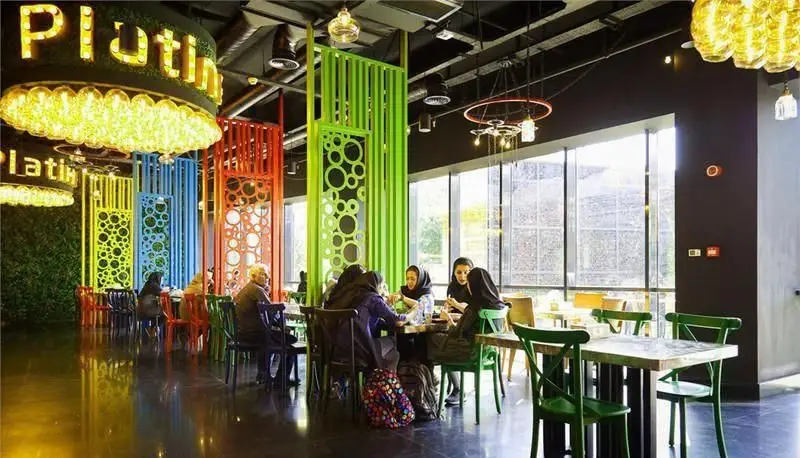 پلاتین فود گاردن: فضای داخلی مدرن و رنگارنگ با پارتیشن‌های تزئینی، میز و صندلی‌های سبز، مشتریان و پنجره‌های بزرگ.