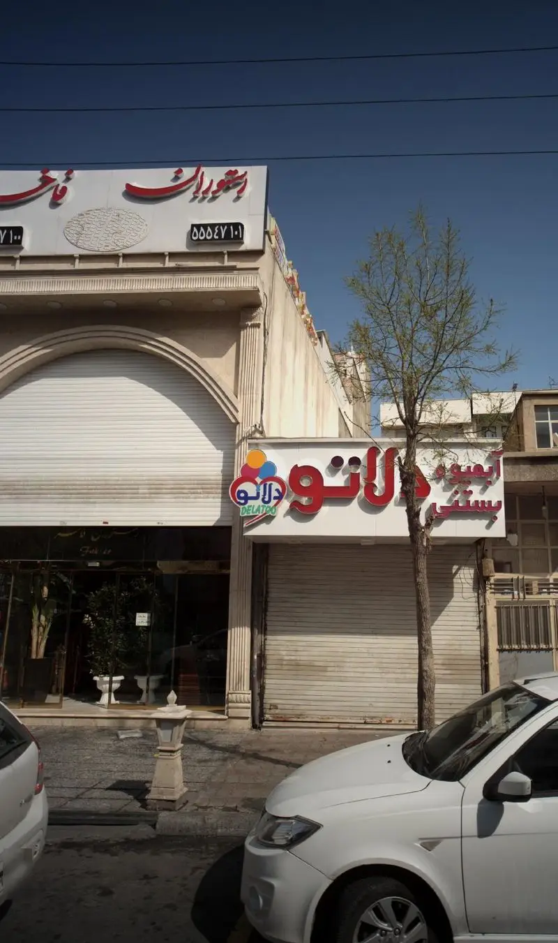 نمای بیرونی رستوران فاخر با تابلوی قرمز رنگ و ساختمان تجاری در روز آفتابی، همراه با تابلوی بستنی دلاتو و ماشین‌های پارک شده.