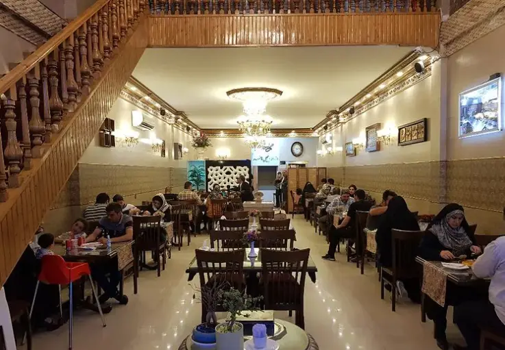 نمای داخلی شیک و دلباز رستوران فاخر: مشتریان در حال صرف غذا در محیطی خانوادگی با دکوراسیون زیبا، میز و صندلی چوبی و پله چوبی چشمگیر.