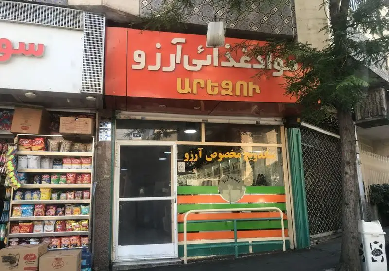 تصویری از نمای بیرونی مغازه ساندویچی و مواد غذایی آرزو با تابلوی پررنگ نارنجی، نوشتار فارسی و ارمنی، و ویترین.