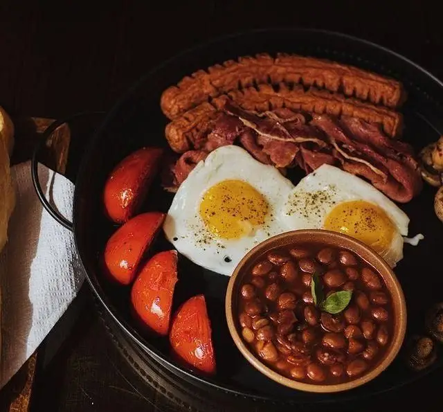 تصویر صبحانه انگلیسی کامل کافه رستوران کاسا دل شف با تخم مرغ، سوسیس، بیکن، گوجه کبابی، لوبیا پخته و قارچ در بشقاب مشکی.