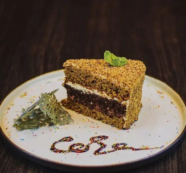 برش کیک چندلایه و خوشمزه با روکش خرده کیک و برگ نعناع، تزئین شده با سس شکلات و اسپرینکل، از منوی دسر کافه رستوران کاسا دل شف.