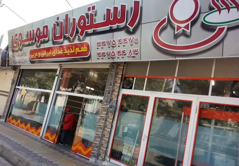 نمای کلی رستوران موسوی با تابلوی بزرگ حاوی نام رستوران، تلفن‌های تماس و شعار 'طعم لذیذ غذای ایرانی و عربی'.