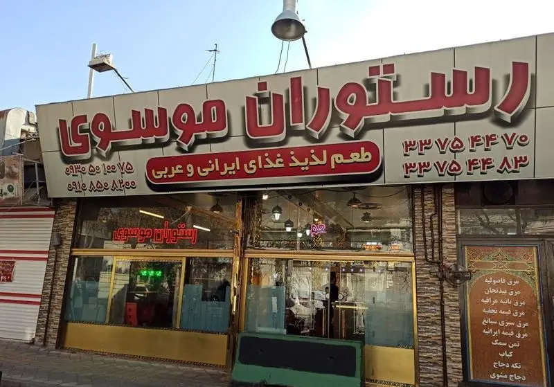 نمای بیرونی رستوران موسوی با تابلوی بزرگ و روشن، ارائه دهنده طعم لذیذ غذاهای ایرانی و عربی، به همراه شماره تماس‌ها و منوی متنوع شامل کباب.
