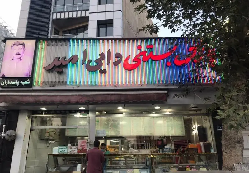 نمای بیرونی مغازه بستنی فروشی دایی امیر در شعبه پاسداران، با تابلوی نئونی رنگارنگ و عکس بنیانگذار. بستنی های سنتی و خوشمزه در تهران.