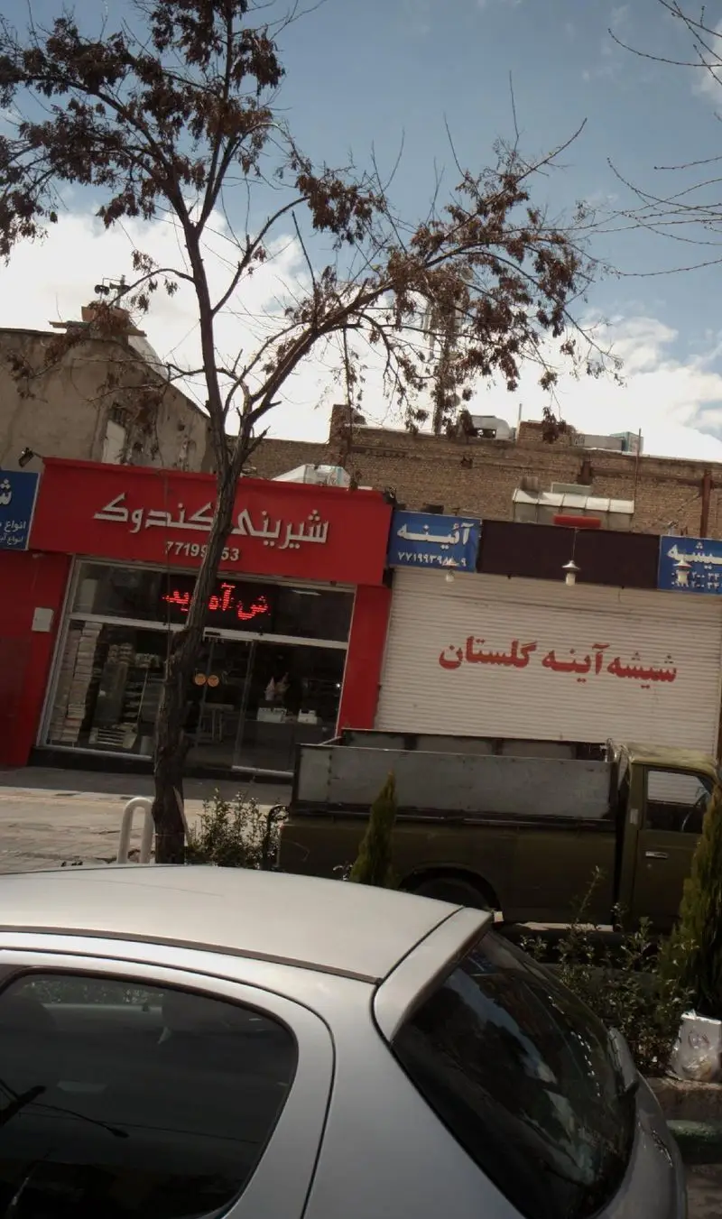 تصویری از نمای بیرونی شیرینی کندوک با سردر قرمز روشن و ویترینهای شلوغ، در کنار تابلوی شیشه آینه گلستان، در خیابانی با درخت و خودرو.