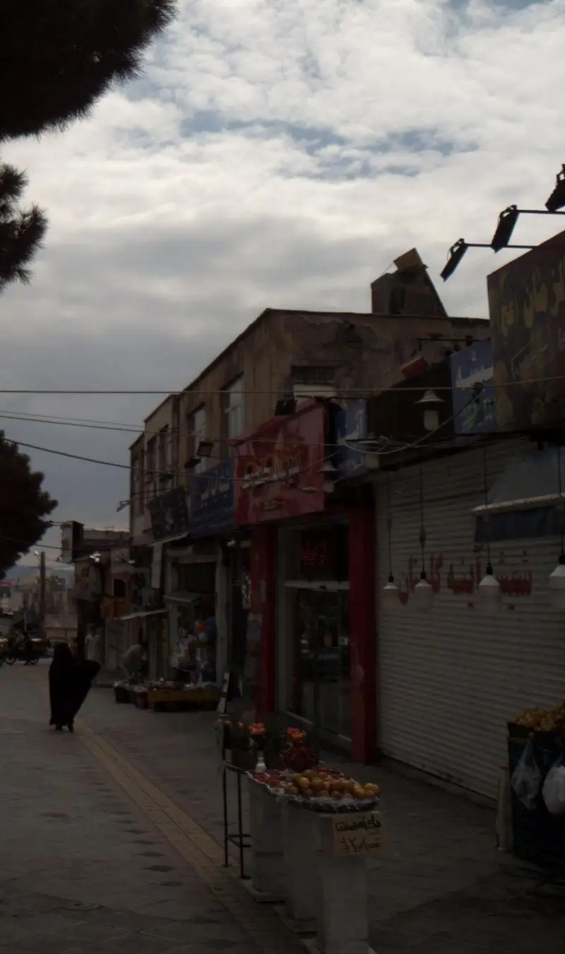 نمایی از خیابان شلوغ با فروشگاه شیرینی کندوک، مردم در حال عبور و مرور، و غرفه میوه. آسمان ابری و حال و هوای بازار محلی.