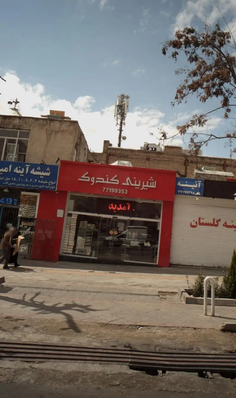 شیرینی کندوک، یک قنادی معروف با تابلوی قرمز رنگ در کنار خیابان، در یک روز آفتابی. نمای شهری با رهگذران و ساختمانهای مجاور. تلفن 77199353.