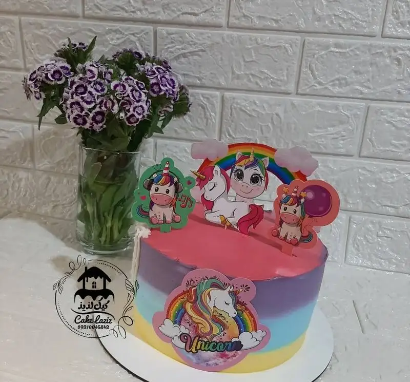 کیک تولد یونیکورن با تزیینات فانتزی و رنگ‌های گرادیانت صورتی، بنفش، آبی و زرد، همراه با تاپرهای یونیکورن و رنگین‌کمان، مناسب جشن کودکان.