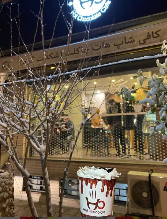 کافی شاپ هات چاکلت در شبی برفی، با تابلوی روشن، درختان پوشیده از برف و لیوان هات چاکلت گرم و لذیذ.
