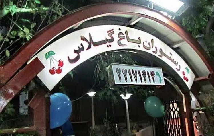 تابلوی ورودی رستوران باغ گیلاس در شب با نورپردازی زیبا، لوگوی گیلاس‌های قرمز و شماره تماس ٢٢١٧١٤١٦. فضایی دلنشین و سبز برای پذیرایی.