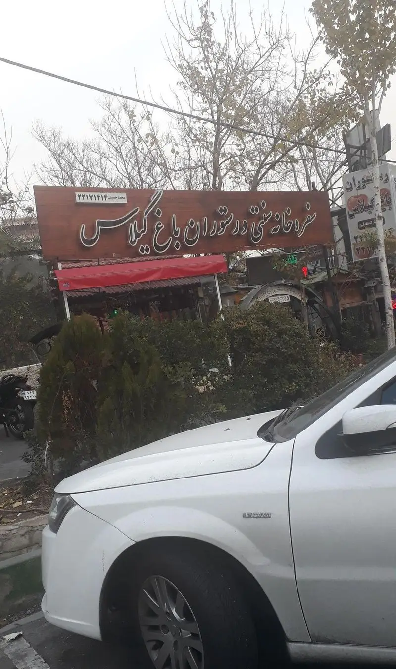 رستوران سنتی و سفره خانه باغ گیلاس با تابلوی چوبی، شماره تلفن ۲۲۱۷۱۴۱۴ و نام محمد حسینی، در فضای سبز کنار خیابان.
