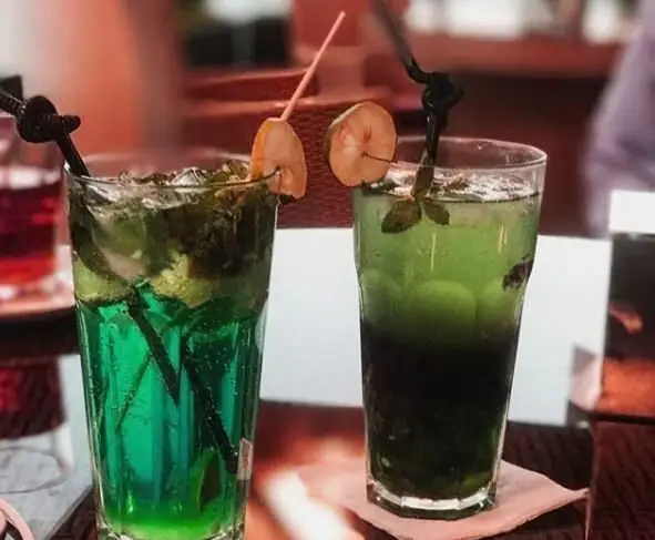 تصویری جذاب از دو نوشیدنی موهیتو سبز و آبی خنک با یخ و نعناع در محیط دلنشین کافی شاپ ایزار. لذت نوشیدنی‌های تابستانی.