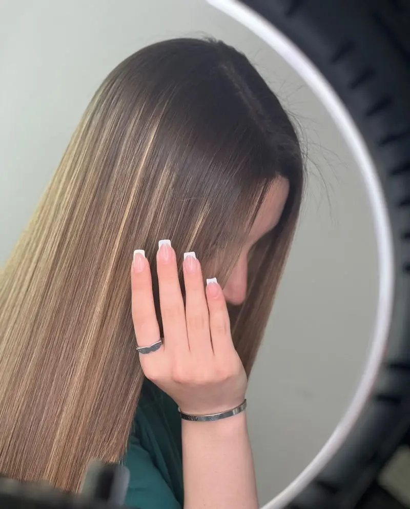 عکس موهای صاف و درخشان با هایلایت عسلی به همراه طراحی ناخن فرنچ سفید و شیک روی دست دختر جوان در سالن زیبایی تخصصی