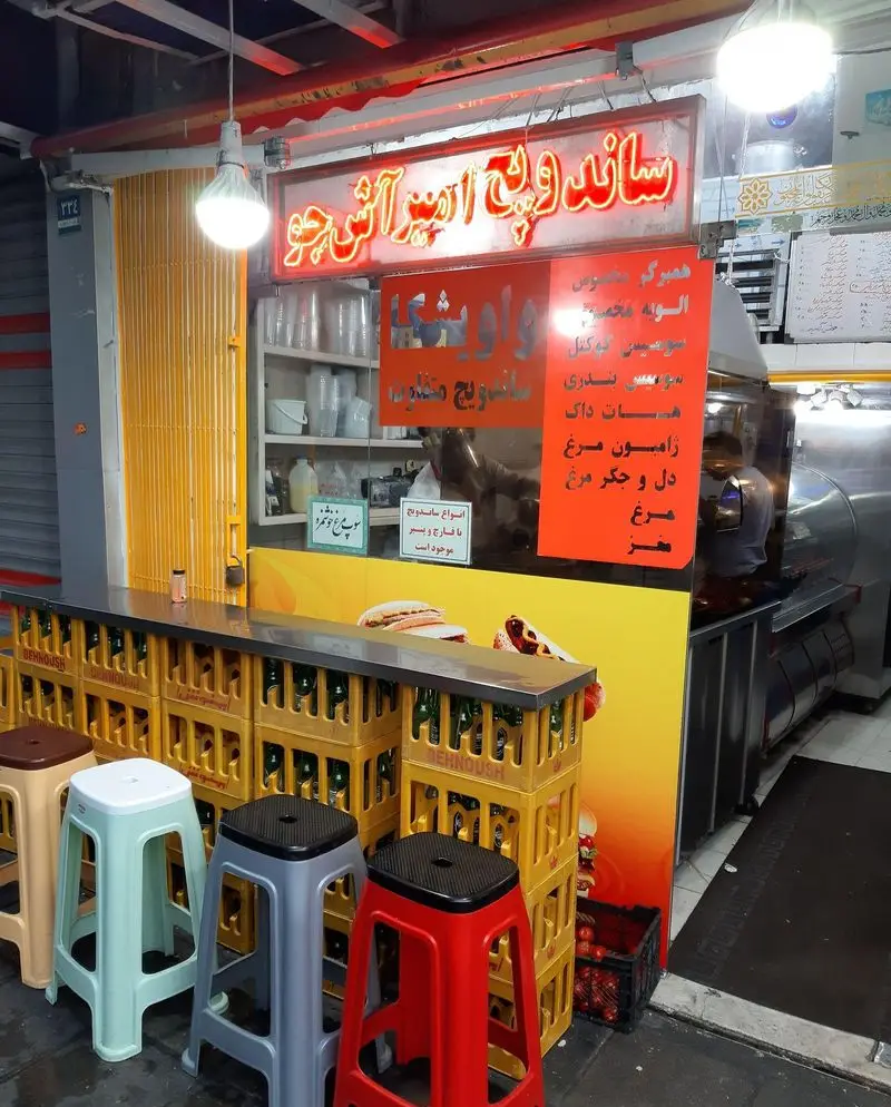 نمای بیرونی ساندویچی امیر با میز ساندویچ‌، صندلی‌ برای مشتریان و منوی کامل انواع ساندویچ‌ها و فست‌فودها در فضای شب.