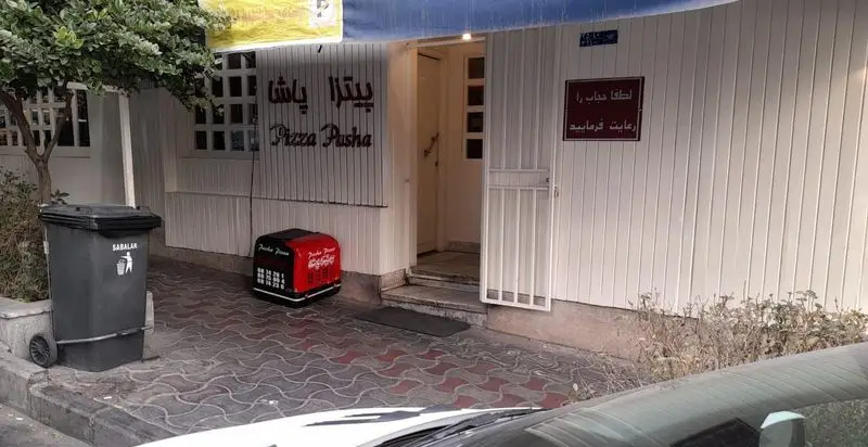 نمای بیرونی فست فود پیتزا پاشا شامل تابلوی رستوران، ورودی اصلی، سطل زباله سبلان و تابلوی رعایت حجاب. جعبه دلیوری پیتزا نیز کنار ورودی دیده می‌شود.