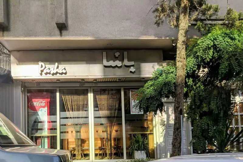 تصویر نمای بیرونی فروشگاه پیتزا پاشا، یک رستوران فست فود محبوب با تابلوی 'Pasha' و 'پاشا'، همراه با یخچال کوکاکولا و دکوراسیون مدرن.