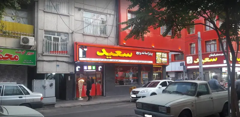 نمایی از فست فود پیتزا و ساندویچ سعید در یک خیابان شلوغ شهری، با تابلوی درخشان قرمز و زرد رنگ و خودروهای پارک شده.