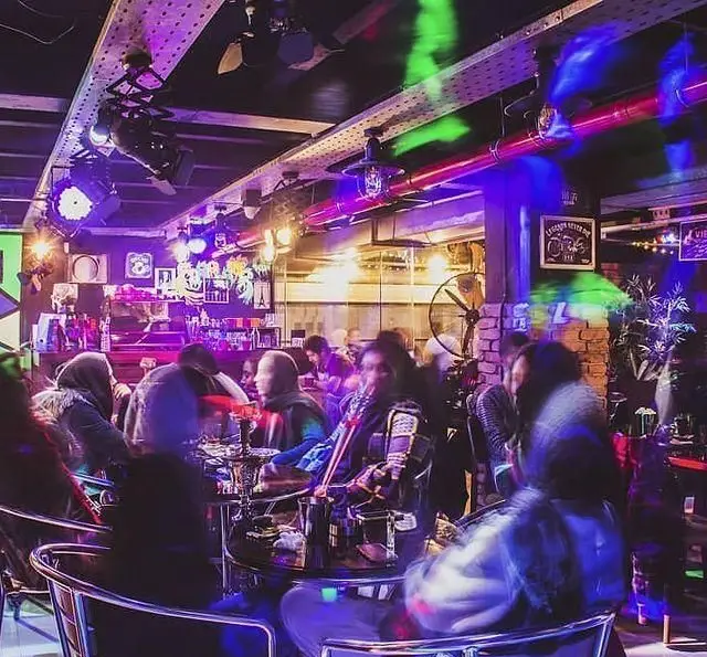 کافه رستوران فروهر: فضای داخلی دنج و مدرن با نورپردازی رنگی، مشتریان در حال لذت بردن از قلیان در محیطی پرجنب و جوش و دلنشین.