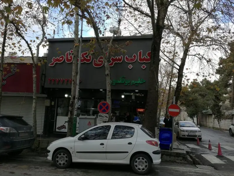 تصویری از نمای بیرونی کباب و حلیم ناصر، رستورانی محبوب با منوی کباب و حلیم، واقع در خیابان شهری با درختان پاییزی و ماشین‌های پارک شده.