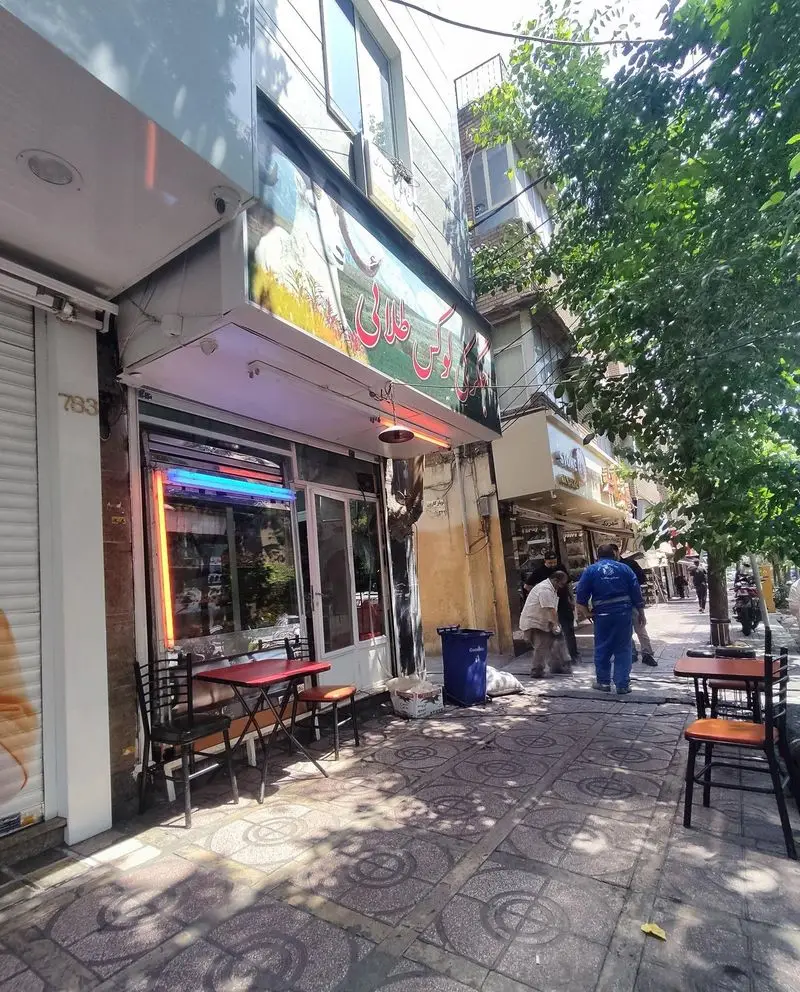 تصویر بیرونی جگرکی لوکس طلائی، رستورانی با میز و صندلی‌های چیده شده روی پیاده‌رو در یک روز روشن و شلوغ شهری.