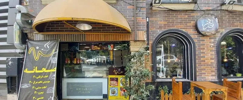 نمای بیرونی کافه و رستوران هتل آپادانا با منوی صبحانه، بستنی ایتالیایی و چیدمان میز و صندلی چوبی در فضای باز