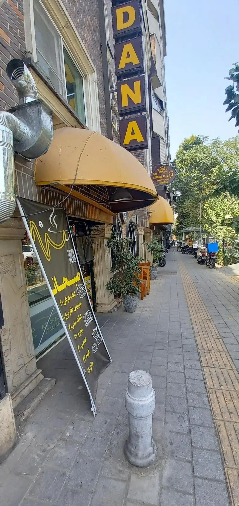 نمای بیرونی رستوران و کافی شاپ هتل آپادانا در خیابانی آفتابی با تابلوهای زیبا، سایبان‌های زرد و منوی صبحانه کنار پیاده‌رو