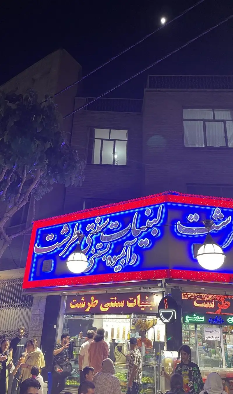 نمای شبانه و شلوغ فروشگاه لبنیات سنتی و آبمیوه بستنی طرشت، با تابلوهای درخشان و جمعی از مشتریان در حال خرید.