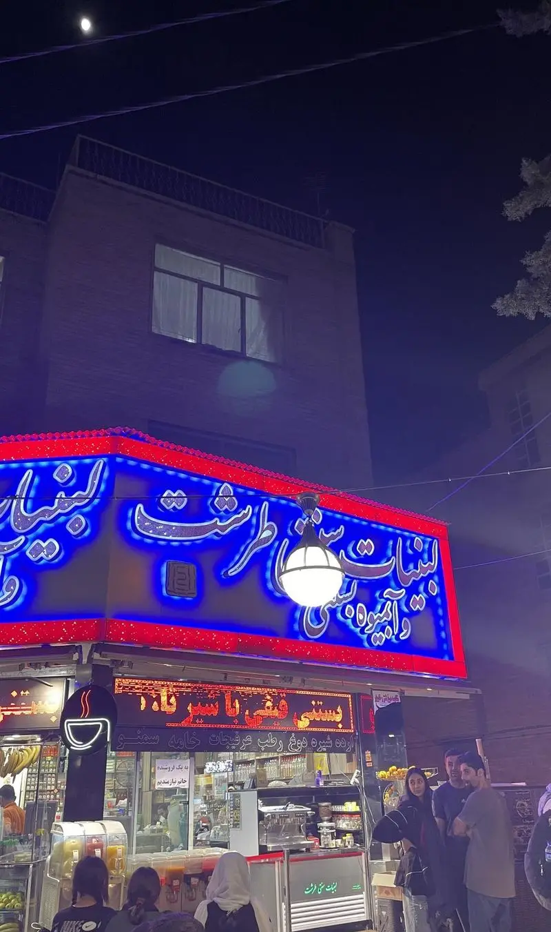 تصویری جذاب از نمای بیرونی مغازه لبنیات سنتی و آبمیوه بستنی طرشت در شب، با تابلو نئون آبی و قرمز و مشتریان.